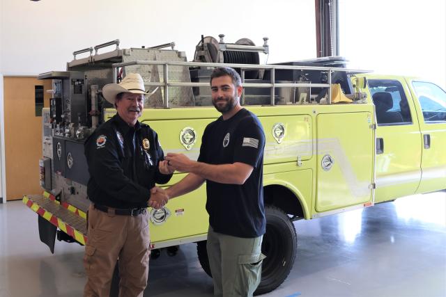 two people shake hands in front of a yellow fire engine. One person hands keys to the other.