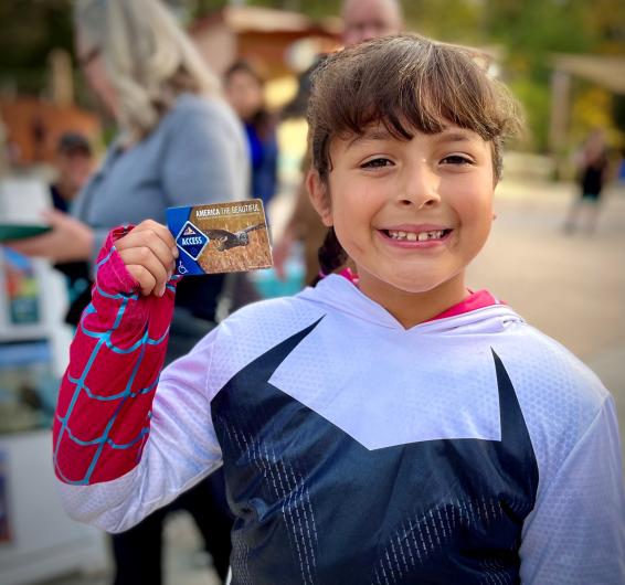 Smiling student, dressed in white and blue, proudly displays her newly acquired Access Pass, which she is holding up. She loves being outdoors and wants to see animals.