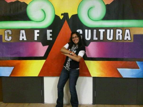 Vanessa Lacayo smiles while standing in front of a stage backdrop with vibrant green, yellow, red, black, and blue geometric designs. The words “Cafe Cultura” are spelled out in white letters in the backdrop.