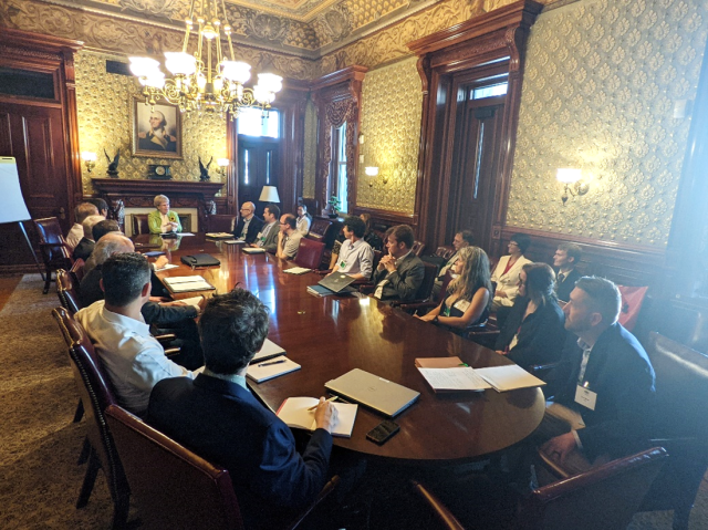 Group of people sit around a table looking at Dr. Jane Lubchenco, located in a large room.
