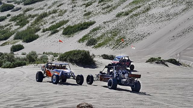 Alt Text: A photo showing three 4-wheeled off-highway vehicles driving on a flat sandy surface. Steep sandy hills with tire tracks and sparse vegetation can be seen in the distance, showing that this is a popular OHV area.