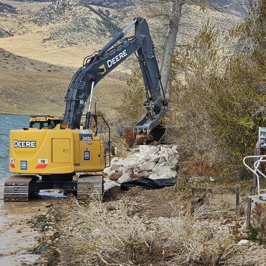 A piece of heavy equipment places large boulders on the shore of a lake.