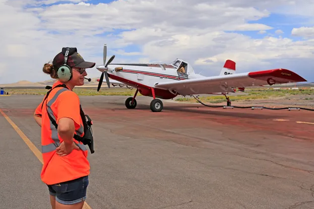 A caucasian woman in a bright orange shirt and headphones stands on weathered tarmac near a small airplane.