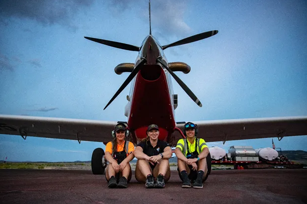 Three Caucasian women sit on the ground in front of a small airplane
