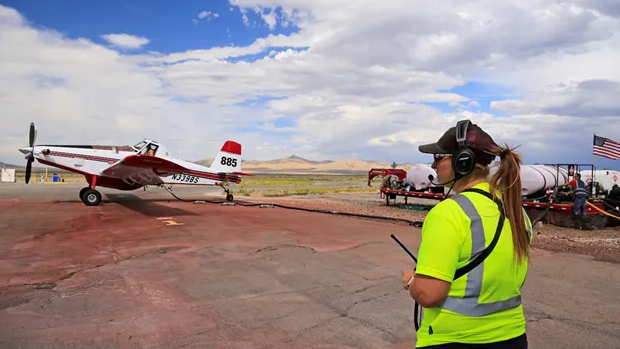 A Caucasian woman in a neon green shirt and headphones stands on weathered tarmac near a small red and white airplane