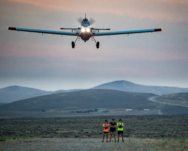 Three people stand close together watching a small plane take off.