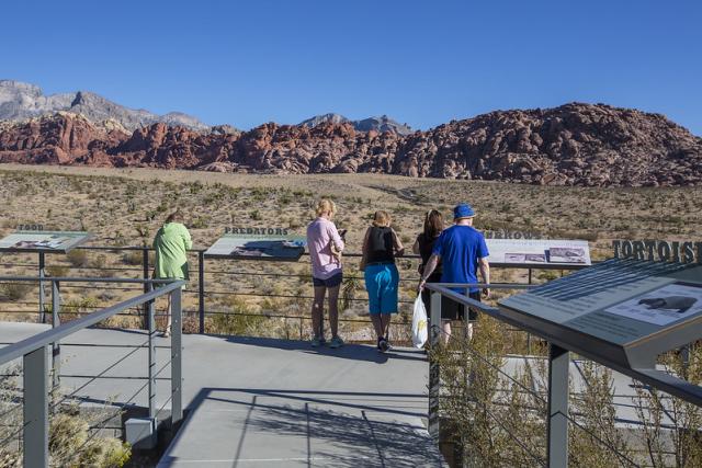 Visitors on a walkway reading panels at chest-height. In the distance are rock formations.