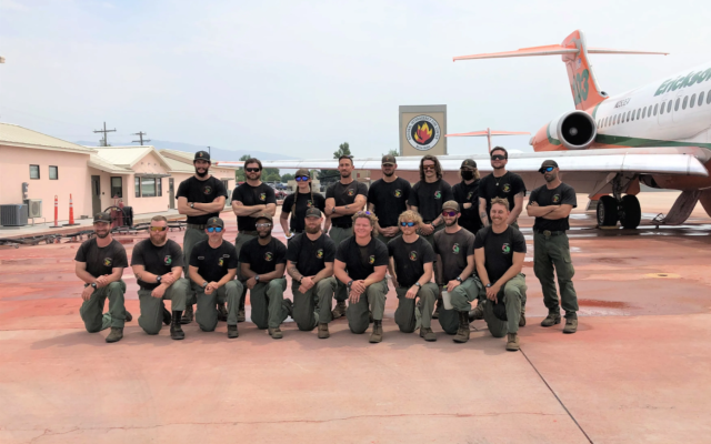 A group of 18 people in green pants and brown tshirts poses in two rows on airplane tarmac.