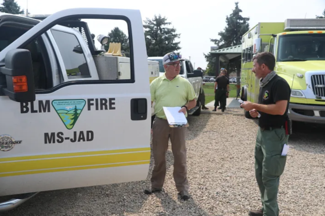 Two caucasian men speak next to an open truck door labeled BLM Fire, MS-JAD.
