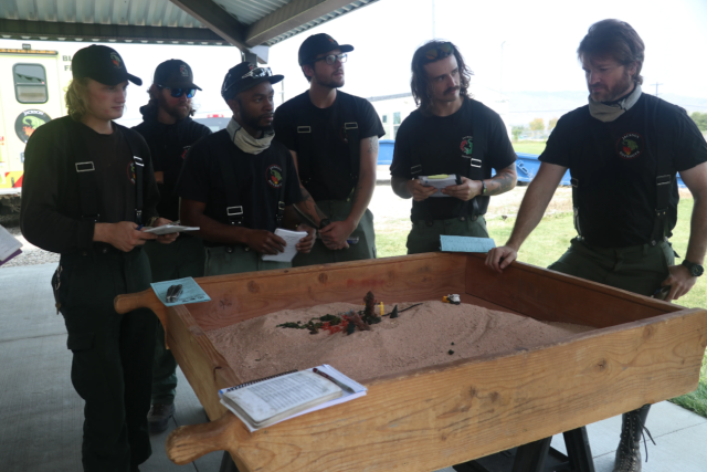 Six young men in tshirts and suspenders stand around a large wooden box filled with sand and toys.