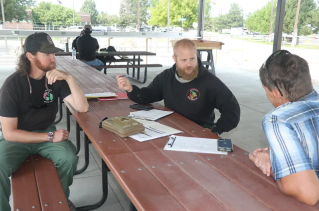 Three caucasian men sit in conversation at a wooden picnic table.