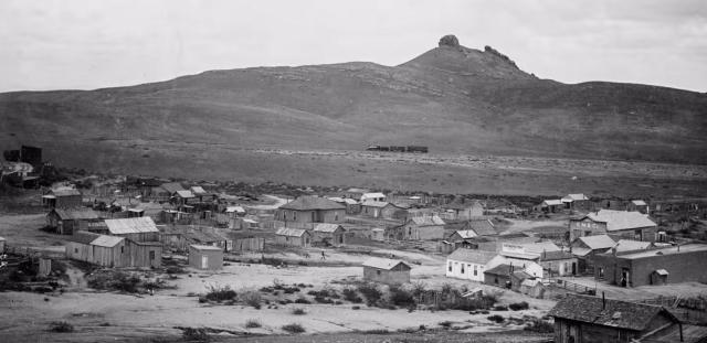 An Old West mining town with wooden buildings in the foreground and a hill in the distance.