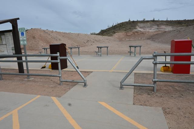A photo showing part of the striped access aisle for the accessible parking spot, with a concrete sidewalk that leads into a wide concrete platform. Straight ahead are three fixed shooting tables. In the foreground is a brown trash bin and a red bin for brass.