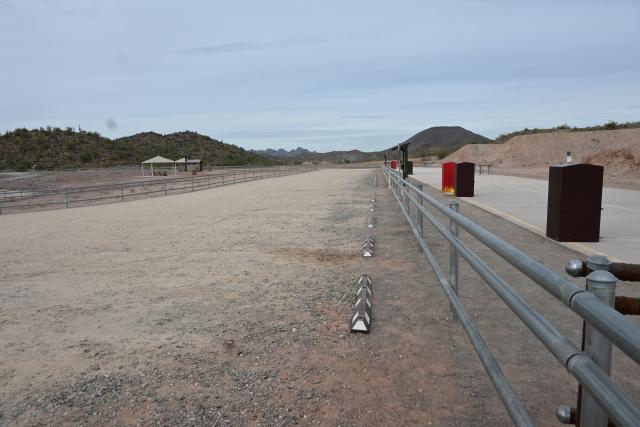 A photo showing a strip of packed gravel, which is a parking lot. On the left and right are metal pole fences separating a picnic area on the left and shooting lanes on the right. Metal trash and brass boxes can be seen on the shooting platform.