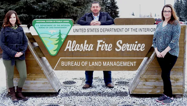 Two Caucasian women and a Caucasian man pose with a sign reading Alaska Fire Service, Bureau of Land Management.