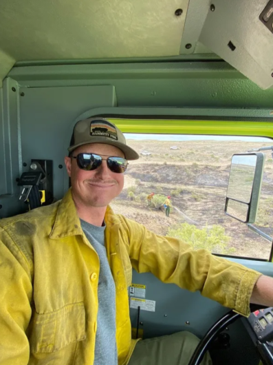 A Caucasian man in a yellow shirt and sunglasses smiles from inside a large truck cab.