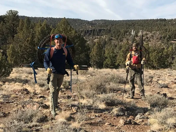 Two Caucasian men in hiking clothes with large rolls of wire and stakes strapped to their backs smiling for the camera outdoors. 