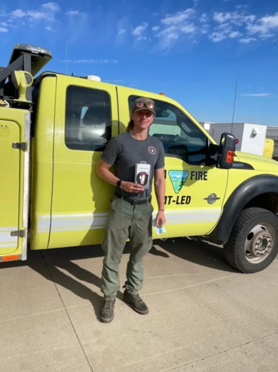 A Caucasian man in a blue tshirt and green pants stands in front of a large bright yellow truck holding up a small box.  