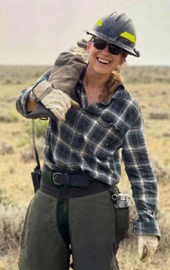A Caucasian woman in sunglasses, a black hard hat, and chainsaw caps smiles outdoors