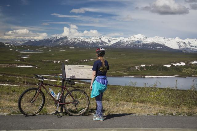 A photo showing a person wearing a bike helmet, standing next to her red bike in front of an interpretive sign that overlooks the Delta Wild and Scenic River. The snow-covered mountains of the Alaska Range can be seen in the background.