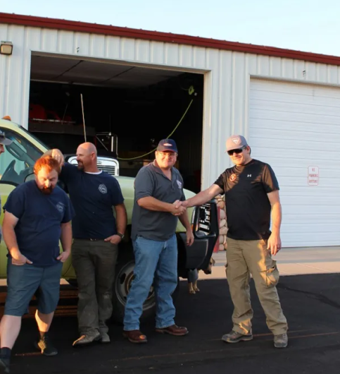 Four caucasian men, two of whom are shaking hands, stand in front of a large truck by a garage