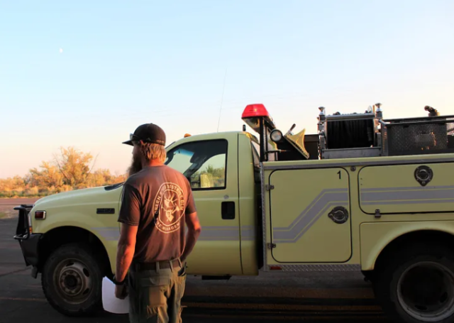 A Caucasian man in a tshirt reading Green River District BLM stands next to a yellow firefighting truck