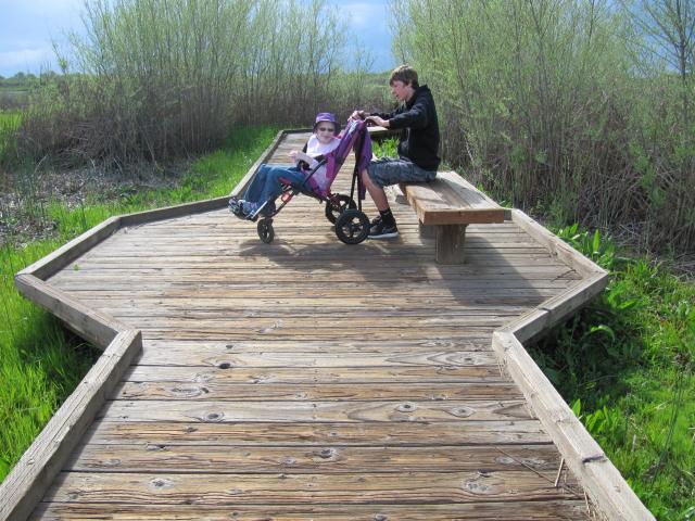 A photo showing a wooden boardwalk leading away from the viewer. A teenage boy sits on a wooden bench on the widened part of the boardwalk with his younger sister who is sitting in a mobility device.