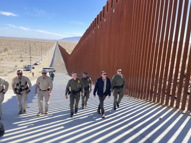 Six people walk along a rust red border wall