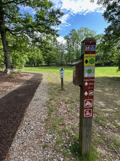 A photo showing a post in the foreground marking the Hidden Pond Trail. It indicates this is an easy, accessible trail that doesn’t allow motorized vehicles, bikes, or horses; and has distances to other trails and features. The trail is on the left, another small sign is a few feet away. Mowed grass and a mixed hardwood forest are in the background.