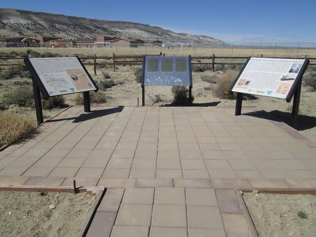 A photo showing a brick path leading to three interpretive signs for the Overland Trail in Wyoming. The signs on the left and right have written descriptions and colorful images and the sign in the middle has three Braille plaques describing the site.