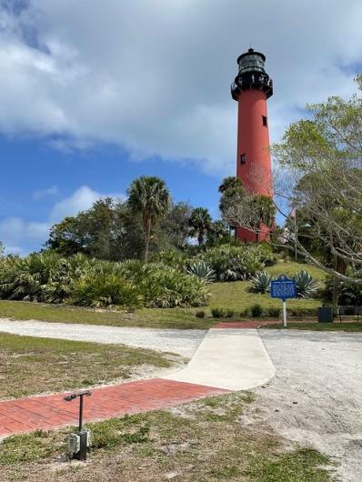 A photo showing a red brick and gray concrete sidewalk over sandy and grassy ground, with a red and black lighthouse in the background on a small hill that’s heavily vegetated with native palm trees and other vegetation.