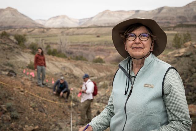 A woman smiles in the front right portion of a photo while scientists in the background conduct a field study of plans along a dirt hillside with mountains in the background