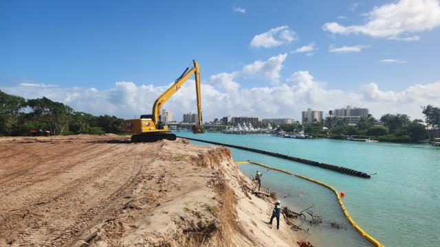 A yellow excavator on a construction site near turquoise waters, with workers and a barrier in the background under a blue sky.