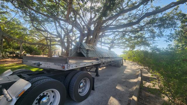 A flatbed truck loaded with stacked metal sheets parked under a large tree in a sunny outdoor setting with a walking path nearby.