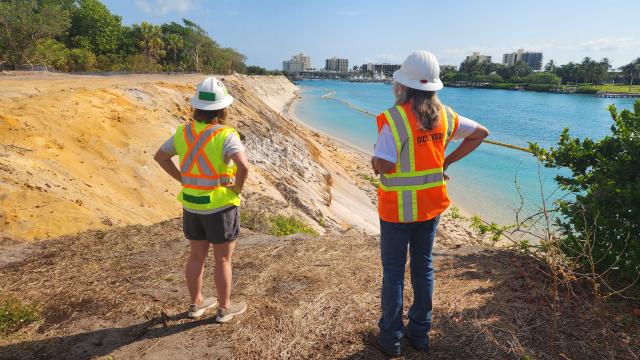 Two women in hard hats and safety vests observe a shoreline project by a clear blue waterway with buildings in the background.