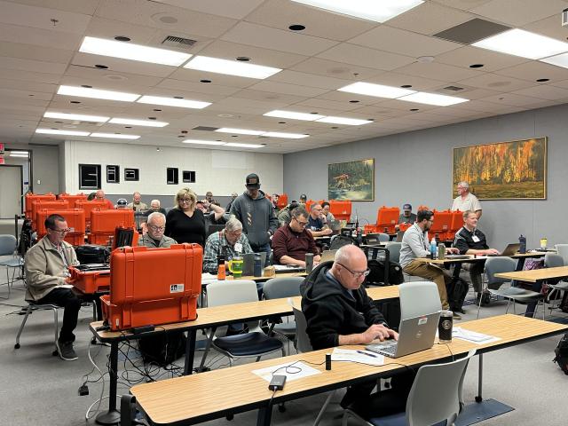 A group of Caucasian men sit indoors at long folding tables