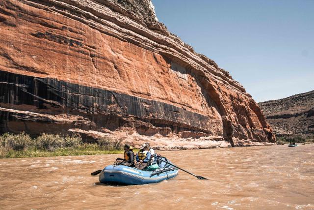 a blue raft with people in it goes down a muddy river with a red and black canyon wall in the background