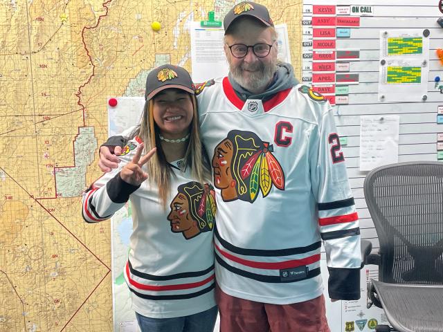  A woman and man stand facing the camera and smiling, wearing matching baseball caps and hockey jerseys to mark Jersey Day at the Dispatch Center. A large map, duty schedule, and notices cover the wall behind them. 