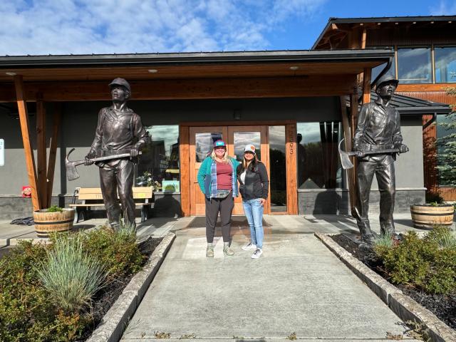  Two women stand facing the camera and smiling near the entrance to a building with an overhang and a doorway framed by brown wooden support beams. They are flanked by two large, dark statues in bronze, which depict firefighters wearing uniforms and protective helmets and each carrying a fire-fighting tool called a Pulaski axe, which has a straight blade on one side and a curved blade, called an adze, on the other.