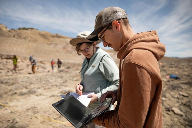 a man holds a laptop while a woman consults a notebook with people looking at a field of dirt and plants in the background