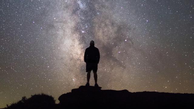 A photo showing the silhouette of a man standing on a high point of ground looking up at the night sky, which is a blend of gray and beige, dotted heavily with stars. The Milky Way is positioned vertically in the center of the photo.  