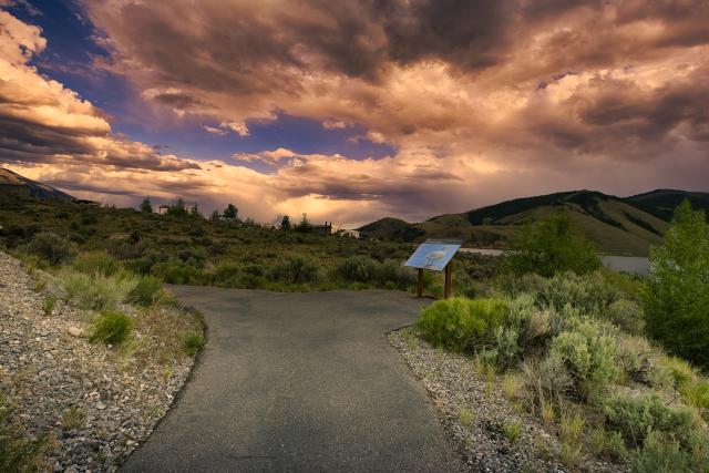 A photo showing an asphalt path leading away from the photographer. A pullout with a colorful sign is in the middle ground on the right. The ground on either side of the path is gravel and heavily vegetated with low shrubs and a few cottonwood trees. 