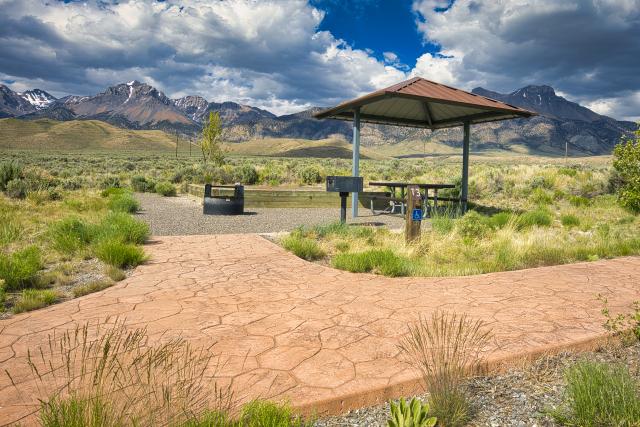 A photo showing a stamped concrete path leading to an accessible campsite with a covered picnic table, grill, fire ring, and elevated tent pad. A mountain range with a dusting of snow is in the distance. Vegetation is a mix of native grasses and sagebrush.  