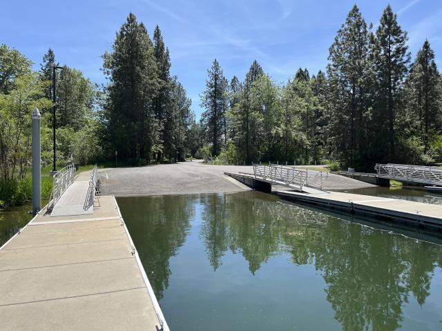 A photo showing a view from a fishing and boat dock in the water looking back at shore. Two wide gravel ramps serve as boat ramps. They go into the water and are flanked by three floating docks that are accessed on shore by ramps with metal railing. Beyond the boat ramps are dense mixed conifer trees. 