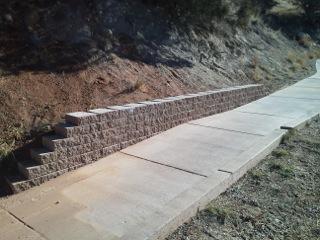 A photo showing a concrete sidewalk with a curb on the right side of it. On the left side of the path is a short brick wall holding back erosion from a short hillside. 