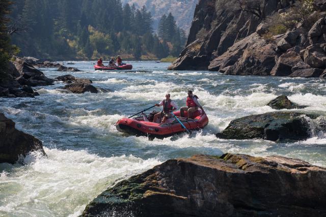 A red raft with two men aboard – both wearing caps, sunglasses, and life jackets – are in the middle of the wide river image. The man in the front with the ores navigates the white water around large boulders while the other man holds onto the raft for stability. A wall of exposed mountain rock is behind them. Another red raft is up the river a few yards in the background with three men aboard. They are paused in calmer water, waiting for the raft in the foreground to clear the gauntlet of white water and r