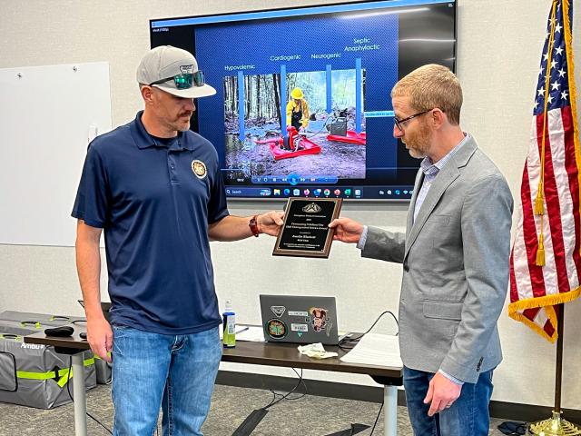 Austin Blackett, left, is handed a plague from Matt Preston, right. Austin is a slimmer, middle aged male with a dark mustache wearing blue jeans. Matt is a slim, middle aged male standing slightly shorter than Austin. Matt is wearing a gray suit jacket with blue jeans.