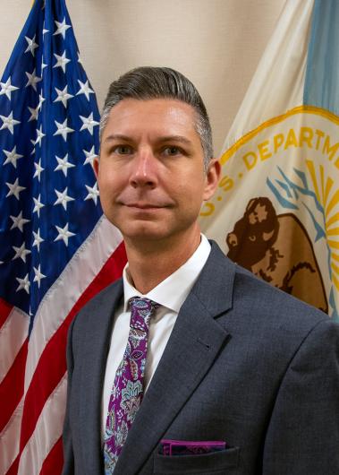 A professionally dressed man in a suit and floral tie stands in front of the American flag and a U.S. Department of the Interior flag. He has short, neatly combed dark hair with some gray, and a neutral expression.