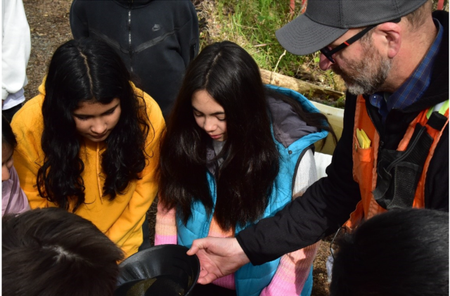 A group of kids crowd around a man wearing a fluorescent safety vest and gray baseball cap. The kids wear brightly colored clothing and each of them are looking down at the black gold pan that the man is holding. The pan is partially filled with water, but a couple of gold flakes are visible at the edge of the pan.