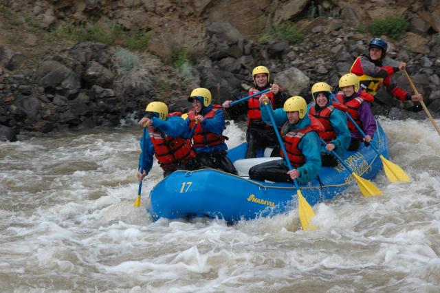 A group of 7 people are in a blue raft with blue and yellow ores making their way through river rapids.